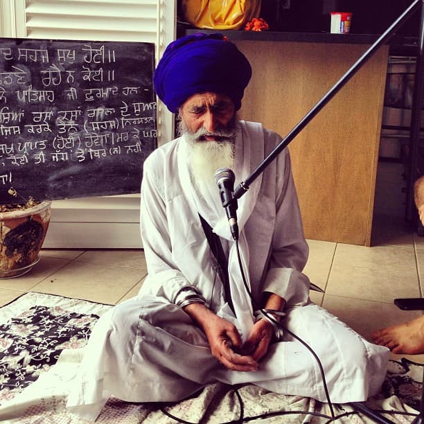 A respected Gurmukh sevadar teaching Gurbani from a chalkboard, in white bana and blue dastaar