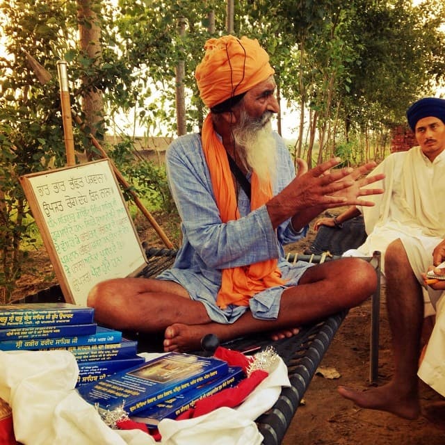 An elder sevadar teaching one-on-one Santhiya outdoors, with a chalkboard and pothis
